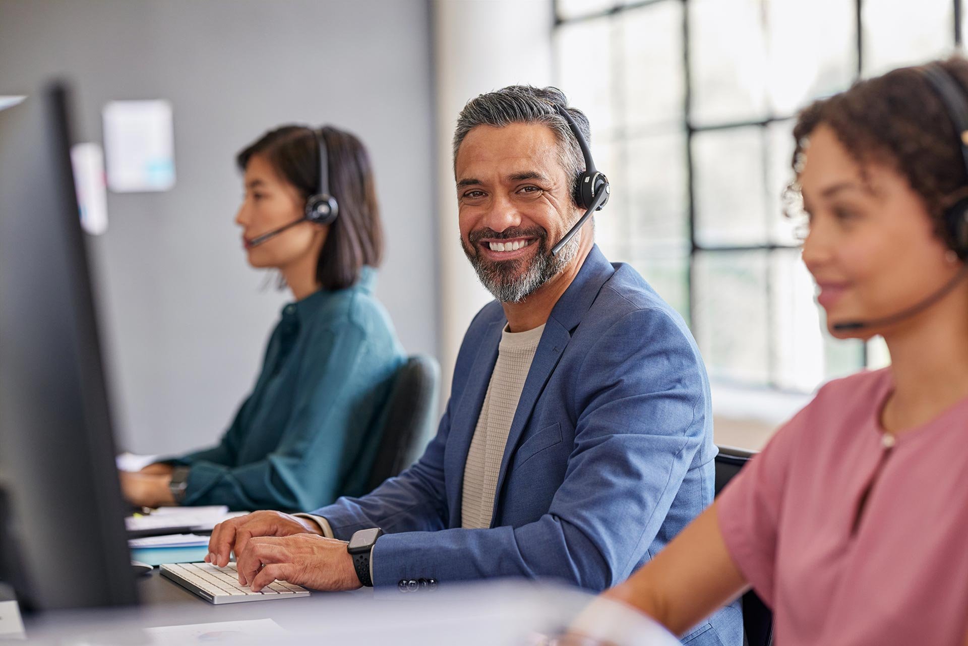 Lächelnder Mann mit Headset im Callcenter, arbeitet am Computer neben Kollegen
